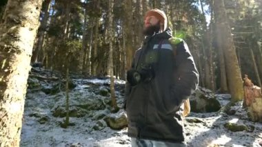 Bir sırt çantası ve arka aydınlatma ve güneş ışığı içinde bir iğne kaplı kış ormanı içinde bir kamera ile ciddi sakallı adam fotoğrafçı portresi