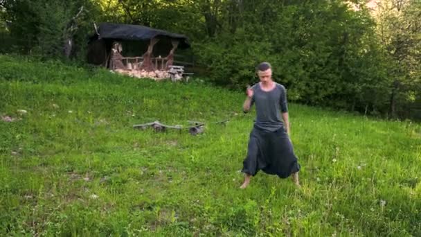 Algemeen plan een jonge man in Indiase kleding en broek Aladdin praktijken Qigong terwijl het doen van oefeningen werken met Qi energie in de zomer buiten naast een oude hut 4k — Stockvideo