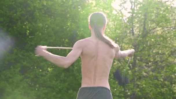 Střední záběr mladý muž s dlouhými vlasy a nahý trup je vycvičen v bojovém umění a předtím, než se v letním ránu vymluví s personálem nebo s lanem v rotaci venku. Nízký úhel — Stock video