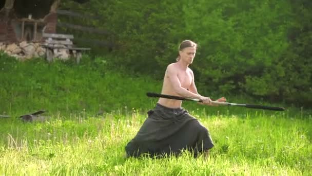 Dlouhý záběr mladý muž s dlouhými vlasy a nahý trup je vycvičen v bojovém umění a předtím, než promluvil s holí nebo holí v letním ránu venku.. — Stock video