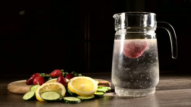 Glazen karaf op een donkere achtergrond met vers fruit en bevroren aardbeien met citroen munt en komkommers in bruisend mineraalwater. Fruit gooien in bruisend water — Stockvideo