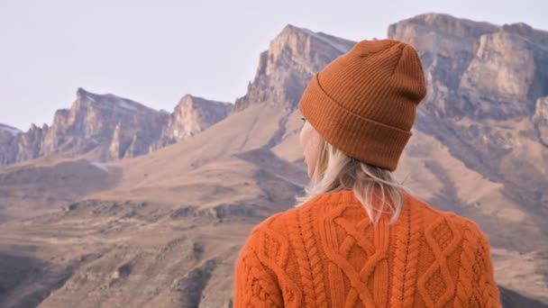 後ろから見る オレンジ色のセーターと帽子をかぶった旅人の女の子が、壮大な山々を背景に立っている。旅行写真家ビデオコンセプト — ストック動画