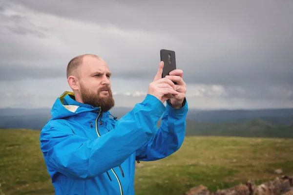 Um homem barbudo tira fotos ou vídeos ao ar livre em seu smartphone. Um cara em um casaco em um fundo de vales de montanha e um céu cinza weedt transmitido pelo telefone — Fotografia de Stock