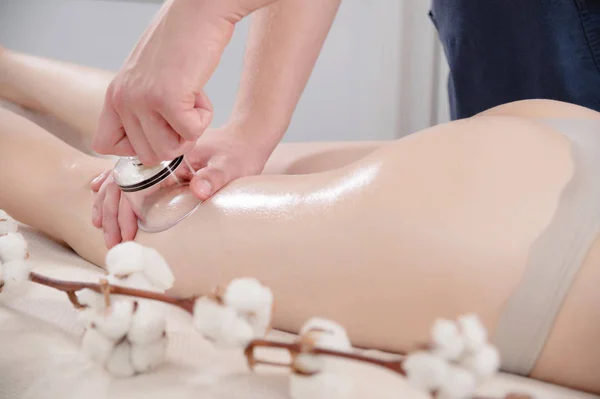 Il primo piano di un fisioterapista maschio che fa un massaggio sottovuoto può massaggiare una ragazza sui fianchi accanto ai fiori di cotone. Dolcezza e attenzione alla salute. Trattamenti cellulite . — Foto Stock