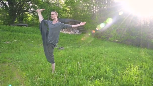 Plano geral um jovem em roupas e calças indianas Aladdin pratica qigong enquanto faz exercícios trabalhando com energia qi no verão ao ar livre ao lado de uma velha cabana 4k — Vídeo de Stock