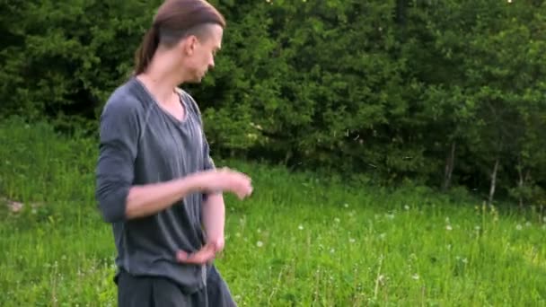 En genomsnittlig plan en ung man i indiska kläder och byxor Aladdin Practices Qigong medan du gör övningar medan du arbetar med Qi energi utomhus på sommaren bredvid en gammal koja — Stockvideo