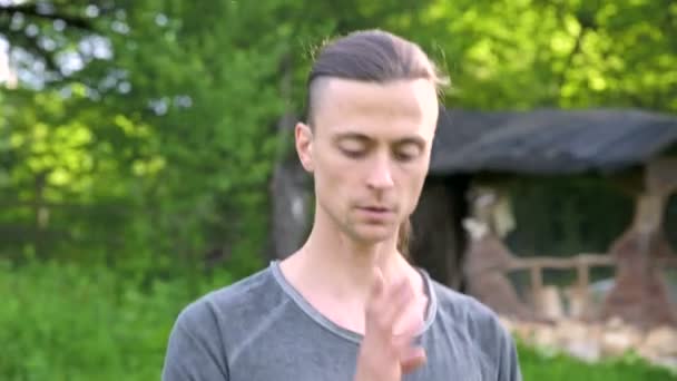 Close-up of a young man in indian clothes and pants alladin practicing qigong while doing exercises while working with qi energy outdoors in summer next to an old hut — Stock Video