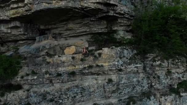 Вид с воздуха на путешественника с рюкзаком, сидящим на краю полки в скале и смотрящим вдаль. Эпическое видео о путешествиях — стоковое видео