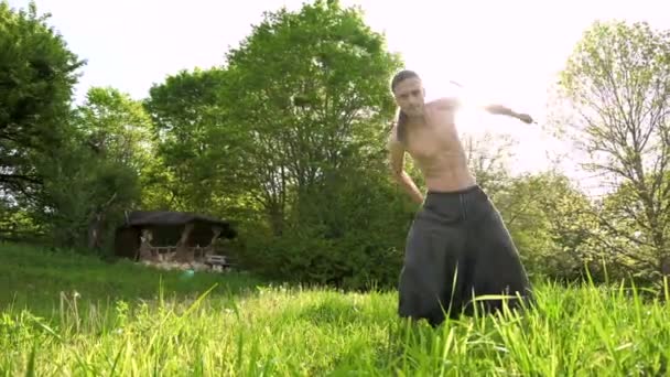 Largo plazo Un joven con el pelo largo y un torso desnudo es entrenado en artes marciales y antes de hablar con un bastón o palo en rotación al aire libre en una mañana de verano. Ángulo bajo — Vídeos de Stock