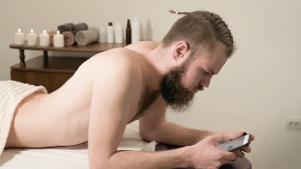 Bearded man with braided hair lying on the medical couch for a massage type SMS message on his smartphone in the office of spa procedures. The concept of freelancing and remote work — Stock Video