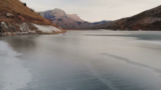 Вид с воздуха на низкий полет над замерзшим озером зимой в окружении Кавказских гор после заката в голубой час — стоковое видео