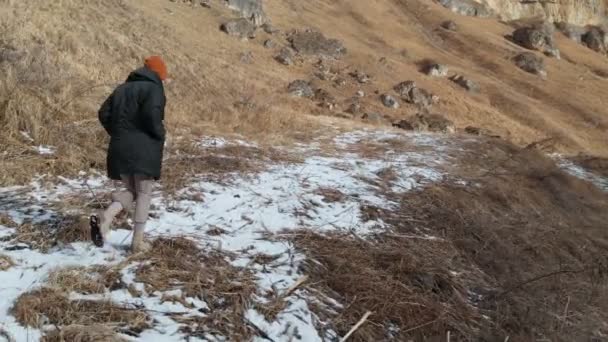 Chica turista en una chaqueta y una gorra camina a lo largo del camino a principios de invierno. hierba amarilla y trozos de nieve en un día soleado. Vista aérea — Vídeos de Stock