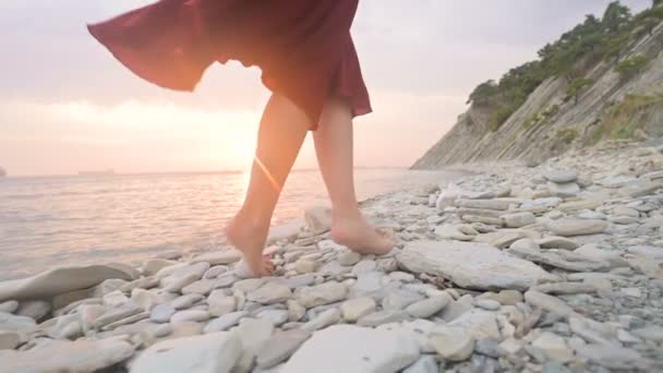 Close-up gerak lambat berikut belakang kaki seorang gadis bertelanjang kaki dalam gaun merah berkibar dalam angin saat matahari terbenam berjalan berjongkok di sepanjang batu pantai laut. Momen ringan dari Tidelessness — Stok Video