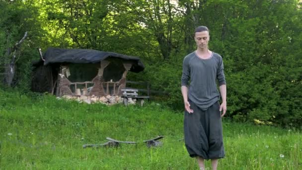 General plan a young man in Indian clothes and pants Aladdin practices qigong while doing exercises working with qi energy in the summer outdoors next to an old hut 4k — Stock Video