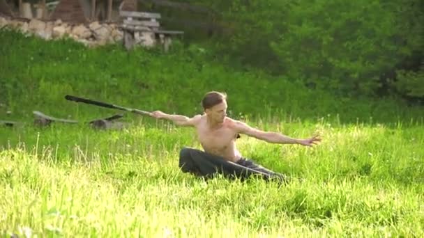 Lang shot een jonge man met lang haar en een naakte torso is opgeleid in Martial Art en voordat spreken met een personeel of stok in rotatie buitenshuis op een zomerse ochtend. — Stockvideo