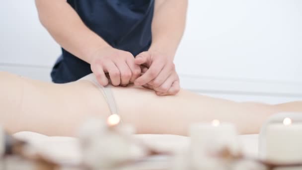 Close-up van een mannelijke fysiotherapeut doet een lichte massage om de heupen en billen van een jonge vrouw op te warmen in een professionele wellnesssalon naast een takje katoen. 4k massage lichaamsverzorging concept — Stockvideo