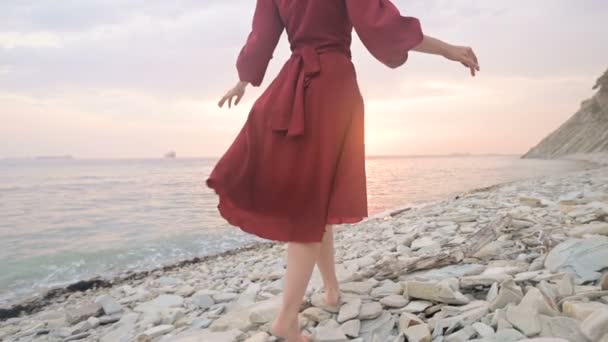 Close-up slow motion following behind the legs of a barefoot girl in a red dress fluttering in the wind at sunset walking crouching along the stones of the sea shore. Light Moments of Tidelessness — Stock Video