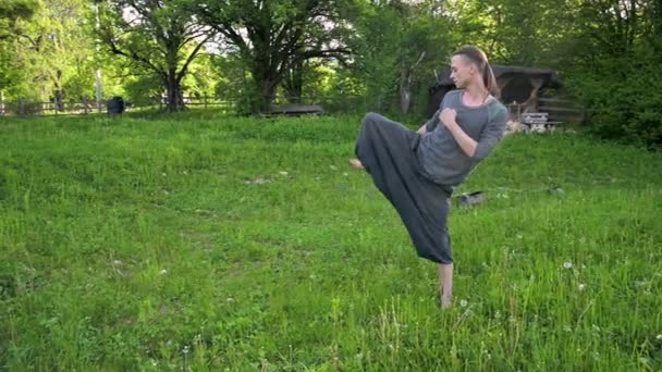 Plano geral um jovem em roupas e calças indianas Aladdin pratica qigong enquanto faz exercícios trabalhando com energia qi no verão ao ar livre ao lado de uma velha cabana 4k — Vídeo de Stock