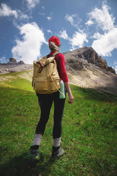 Вид зі спини дівчина спортсмена в червоних сонцезахисних окулярів і жовтий рюкзак стоїть на зеленому схилі на тлі епічних скель Кавказу — стокове фото