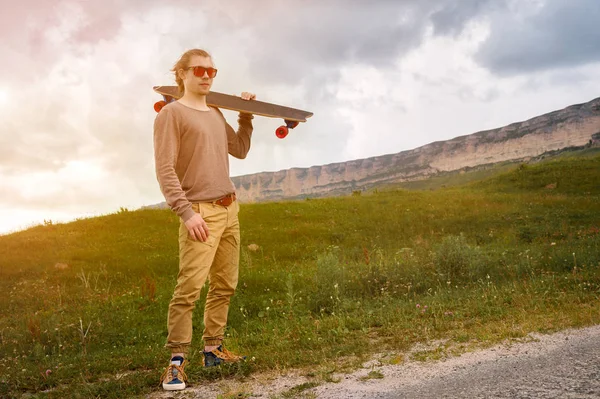 Şık genç adam gün batımından sonra akşam omzunda bir paten veya longboard ile dolambaçlı bir dağ yolu boyunca ayakta. Gençlik spor ve seyahat hobileri kavramı — Stok fotoğraf