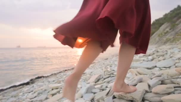 赤いドレスを着た裸足の女の子の足の後ろに続くクローズアップスローモーションは、海辺の石に沿ってしゃがんで夕暮れ時に風に揺れる。潮の流れの光の瞬間 — ストック動画