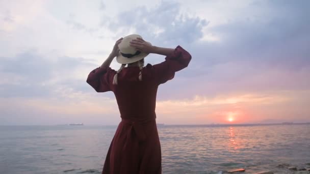 La vista desde atrás. Retrato en cámara lenta de una joven con un vestido rojo y un sombrero de paja en la orilla del mar contra una costa rocosa y puesta de sol rosa.. Esperando el regreso de los marineros — Vídeos de Stock