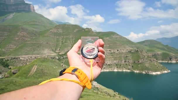 Pandangan orang pertama dari tangan laki-laki memegang kompas magnetik plastik berputar di samping dan mencari arah yang tepat di latar belakang gunung. Konsep orienteering — Stok Video