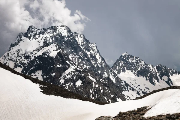 Uitzicht op een hoge steile rotsachtige berg gedeeltelijk bedekt met sneeuw tegen een donkere bewolkte hemel met wolken vastklampen aan de top. Berg weer Verander concept — Stockfoto