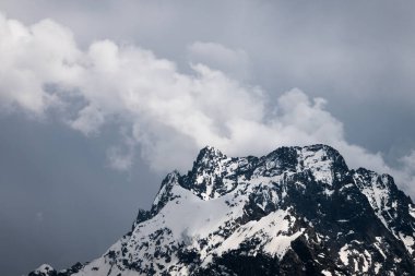 Bulutlar tepeye sarılmış karanlık bulutlu gökyüzüne karşı kısmen karla kaplı yüksek bir dik kayalık dağ görünümü. Dağ hava sıcamı kavramı