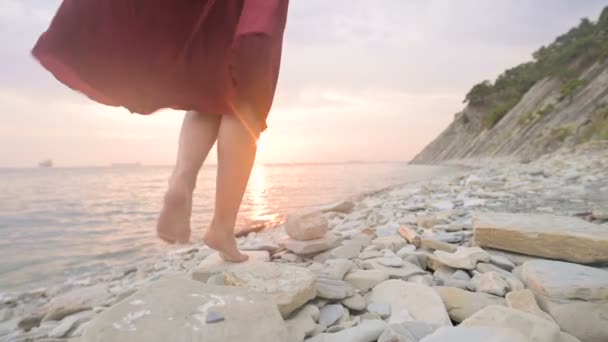 Close-up slow motion following behind the legs of a barefoot girl in a red dress fluttering in the wind at sunset walking crouching along the stones of the sea shore. Light Moments of Tidelessness — Stock Video