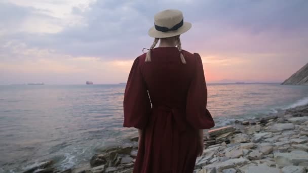 La vista desde atrás. Retrato en cámara lenta de una joven con un vestido rojo y un sombrero de paja en la orilla del mar contra una costa rocosa y puesta de sol rosa.. Esperando el regreso de los marineros — Vídeos de Stock