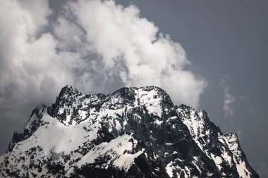 Bulutlar tepeye sarılmış karanlık bulutlu gökyüzüne karşı kısmen karla kaplı yüksek bir dik kayalık dağ görünümü. Dağ hava sıcamı kavramı