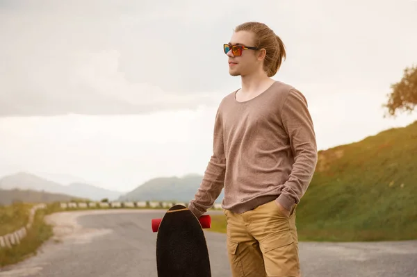Jovem elegante de pé ao longo de uma estrada de montanha sinuosa com um skate ou longboard em seu ombro à noite após o pôr do sol. O conceito de esportes juvenis e hobbies de viagem — Fotografia de Stock