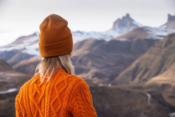 Retrato da parte de trás da menina viajante em uma camisola laranja e chapéu nas montanhas contra o fundo de uma montanha congelada. Foto conceito de viagem — Fotografia de Stock