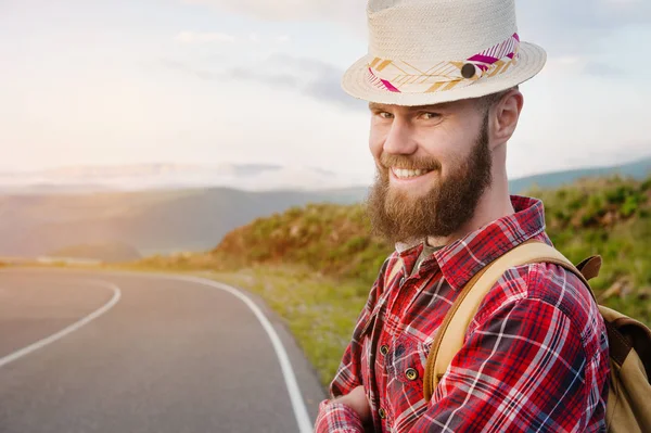 Retrato de um viajante sorridente feliz barbudo hipster com uma mochila em uma camisa xadrez e um chapéu ao lado de um carro desconhecido fica na estrada ao pôr do sol nas montanhas. Conceito de viagem feliz e confiante — Fotografia de Stock