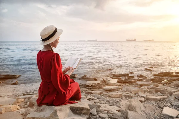 Приваблива кавказька дівчина в червоній яскравій сукні і солом'яний капелюх сидить на великому камені біля моря на заході і читає цікаву книгу про морську горизонт. Перегляд назад — стокове фото