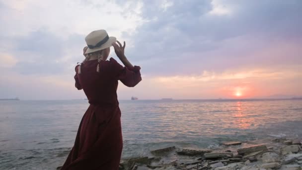 Udsigten fra bagsiden. Langsom bevægelse Portræt af en ung pige i en rød kjole og en stråhat på stranden mod en klippefyldt kyst og lyserød solnedgang. Venter på tilbagelevering af sejlere – Stock-video