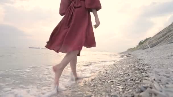 Las piernas de una joven con un vestido rojo por detrás caminando en una playa rocosa la costa del mar al atardecer. Un vestido revoloteando al viento en el viento . — Vídeo de stock