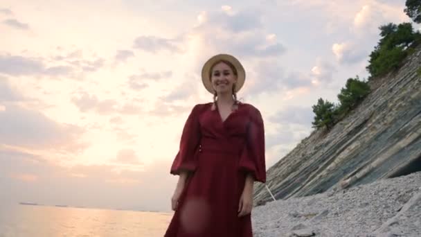 赤い光のドレスと麦わら帽子をかぶった若い魅力的な笑顔の女の子は、日没時の風の強い天候の中、海岸に沿って歩きます。明るさと美しさにおける女性らしさと自立 — ストック動画
