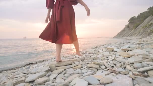 Close-up slow motion following behind the legs of a barefoot girl in a red dress fluttering in the wind at sunset walking crouching along the stones of the sea shore. Light Moments of Tidelessness — Stock Video