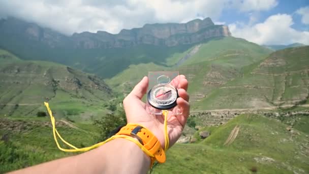 La vista en primera persona de una mano masculina sosteniendo una brújula magnética de plástico gira en el lado y busca la dirección correcta en el fondo de una montaña. El concepto de orientación — Vídeos de Stock