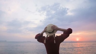 Arkadan manzara. Yavaş çekim Kırmızı elbiseli ve deniz kıyısında hasır şapkalı genç bir kızın portresi. Rüzgardaki bir kız elleriyle şapka tutar. Denizcilerin dönüşünü bekliyoruz