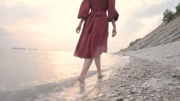 Benen van een jong meisje met een rode jurk van achteren wandelen op een rotsachtig strand de zee kust bij zonsondergang. Een jurk fladderende in de wind in de wind. — Stockvideo