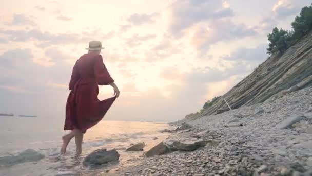 Uma jovem garota atraente em um vestido de luz vermelha e um chapéu de palha caminha ao longo da costa em tempo ventoso ao pôr do sol. Femininidade e independência em leveza e beleza — Vídeo de Stock