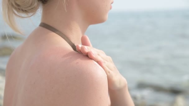 Una joven atractiva que se aplica protector solar en el hombro mientras está sentada en la orilla del mar en un día soleado de verano. Protección de verano y concepto de cuidado de la piel . — Vídeos de Stock