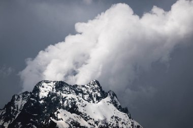 Bulutlar tepeye sarılmış karanlık bulutlu gökyüzüne karşı kısmen karla kaplı yüksek bir dik kayalık dağ görünümü. Dağ hava sıcamı kavramı