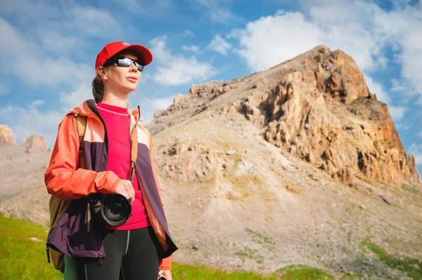 Perempuan atlet fotografer dengan topi merah kacamata hitam dan ransel kuning berdiri di lereng hijau dengan latar belakang tebing epik Kaukasus — Stok Foto