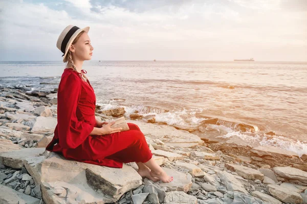 Приваблива кавказька дівчина в червоній яскравій сукні і солом'яний капелюх сидить на великому камені біля моря на заході і читає цікаву книгу про морську горизонт і прекрасні хмари. — стокове фото