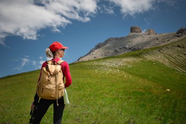 Kırmızı şapkalı güneş gözlüğü ve sarı sırt çantası ile arka Kız atlet görünümü Kafkasya'nın epik kayalıkların arka plan karşı yeşil bir yamaç üzerinde duruyor