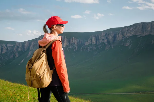 Lány sportoló egy piros sapka napszemüveg és egy sárga hátizsák áll egy zöld lejtőn a háttérben az epikus sziklák a Kaukázusban — Stock Fotó